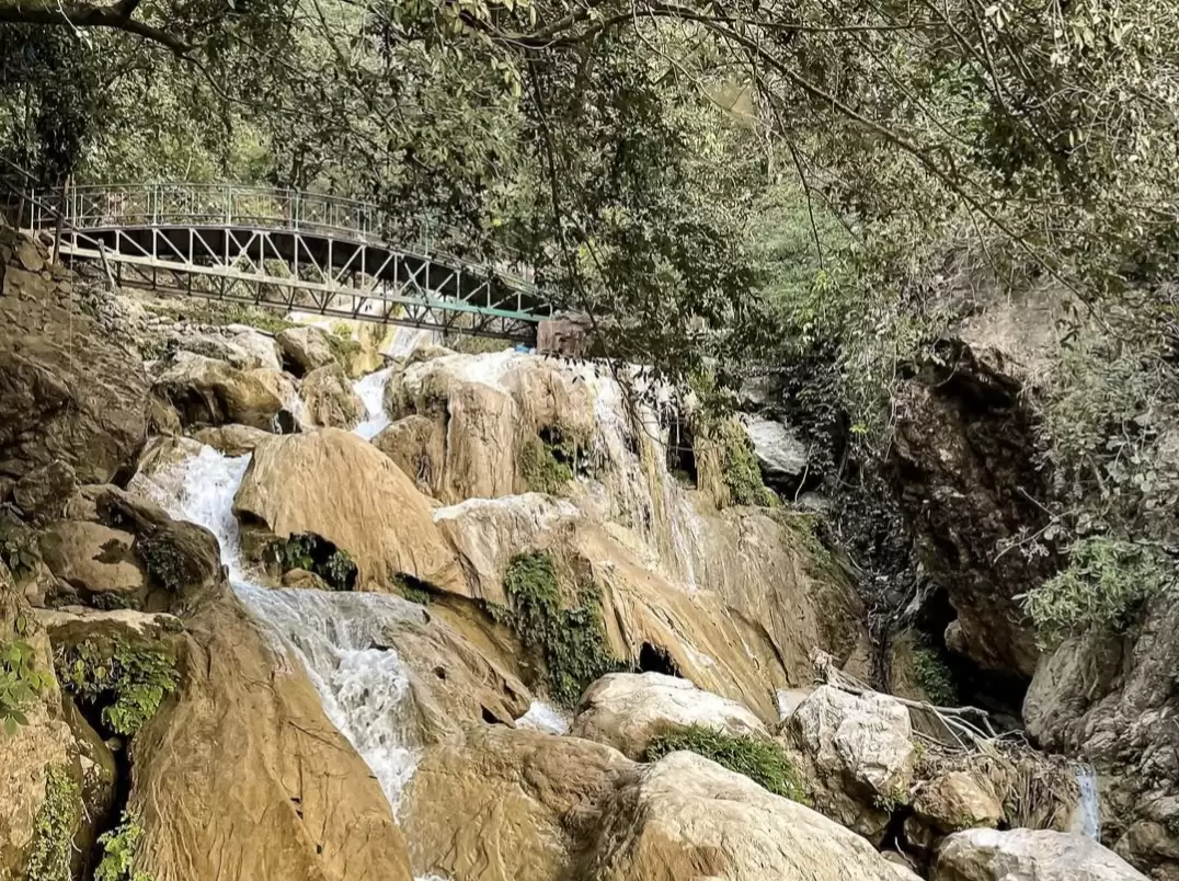 Neer Waterfall at Rishikesh during sunny day, featuring terraced golden cascades iron bridge lush forest greenery rocks, perfect adventure experience Uttarakhand tour package.