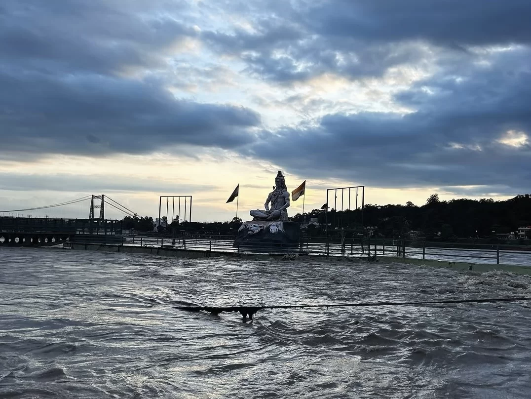 Panoramic view colossal Shiva statue with fluttering saffron flags overlooking swirling Ganga River Rishikesh during dramatic cloudy sunset, suspension bridge poles distant hills townscape water flow railing foreground, perfect Uttarakhand tour package.
