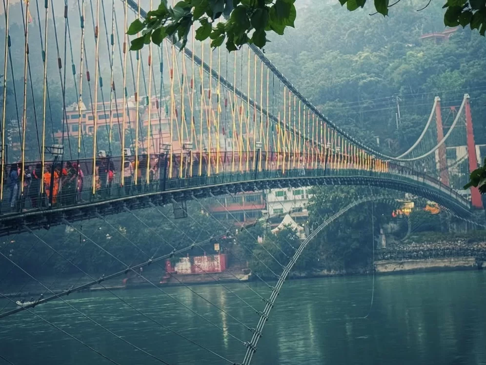 Laxman Jhula suspension bridge at Rishikesh during foggy morning, featuring yellow cables temple buildings Ganga River trees walkers, perfect spiritual experience Uttarakhand tour package.