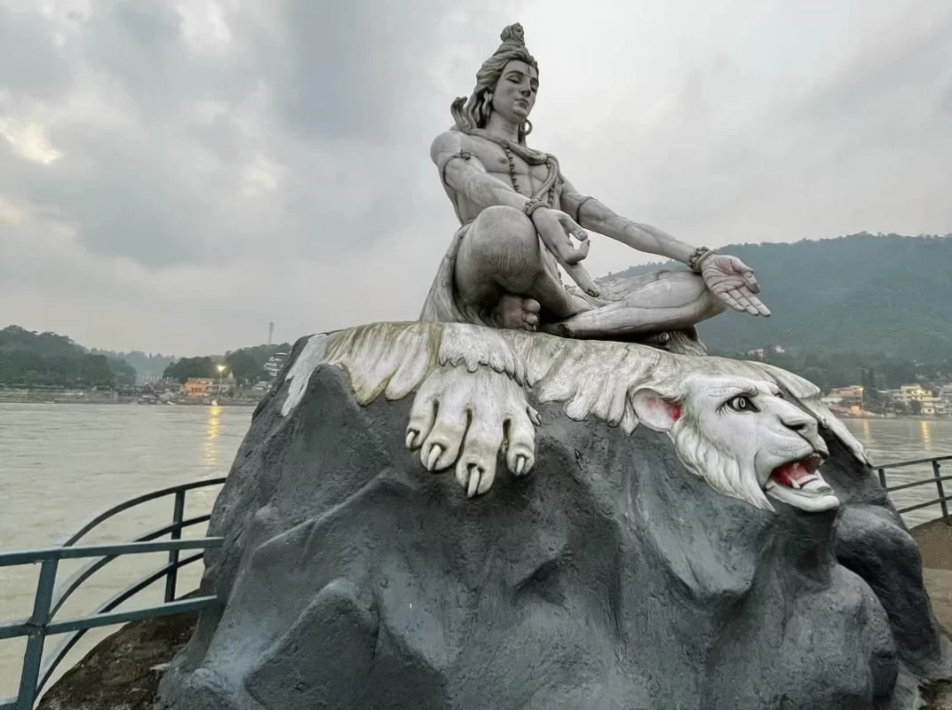 Grand Shiva statue seated meditative pose on roaring lion rock pedestal overlooking Ganga River Rishikesh during cloudy evening, misty hills riverside railing buildings backdrop, perfect iconic spiritual landmark Uttarakhand tour package.