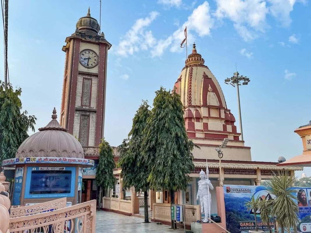 Triveni Ghat temple clock tower at Rishikesh during sunny day, featuring pink domes trees flags statue palms, perfect spiritual experience Uttarakhand tour package.
