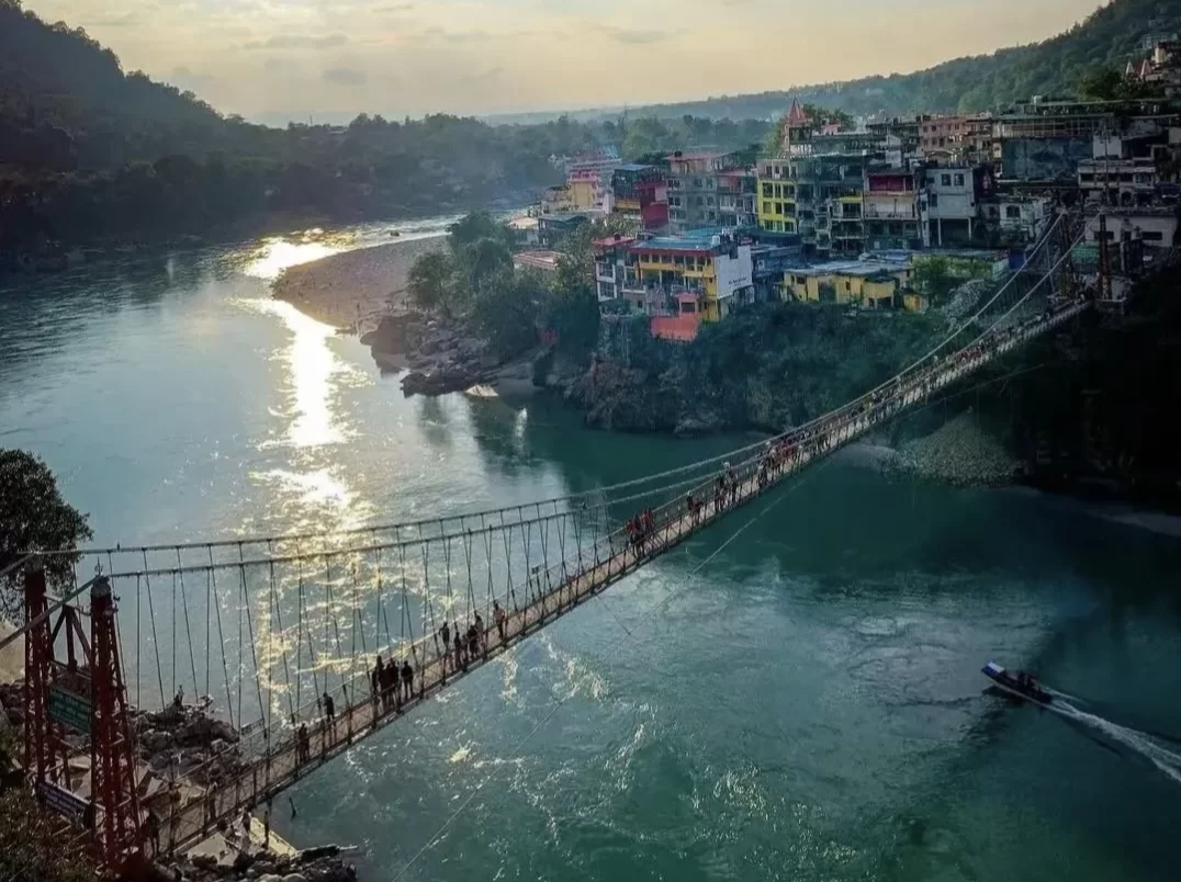 Laxman Jhula suspension bridge at Rishikesh during golden hour, featuring turquoise Ganga River colorful riverside buildings walkers boat hills, perfect spiritual experience Uttarakhand tour package.