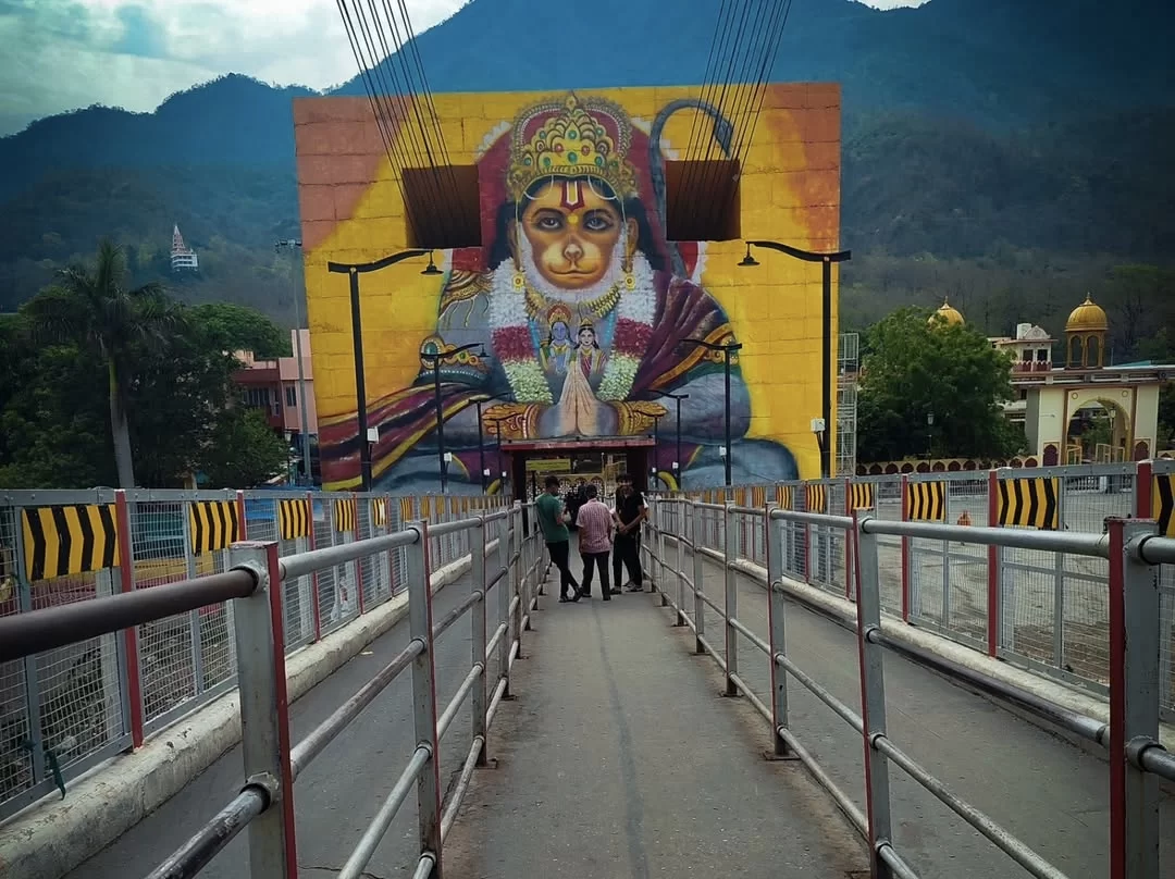 Janki Setu suspension bridge in Rishikesh featuring giant Lord Hanuman mural yellow background praying pose, pedestrians crossing railings Ganges river hills temples backdrop, perfect spiritual experience Uttarakhand tour package.