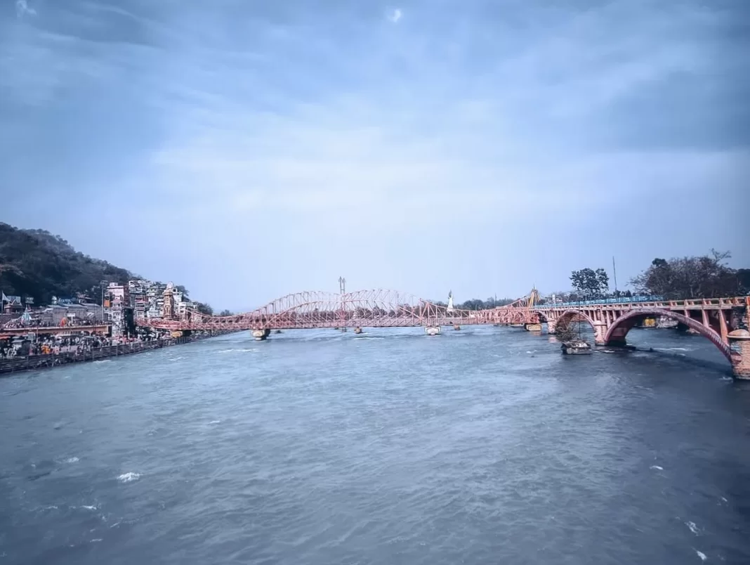 Har Ki Pauri Haridwar iconic pink Lakshman Jhula suspension bridge over Ganga river ghats temples boats cloudy sky hills backdrop, perfect spiritual riverside icon, Uttarakhand tour packages.