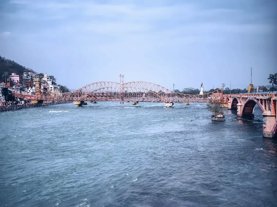 Har Ki Pauri Haridwar iconic pink arched Lakshman Jhula bridge over Ganga river temples boats hills cloudy sky, perfect pilgrimage riverside view, Uttarakhand tour packages.