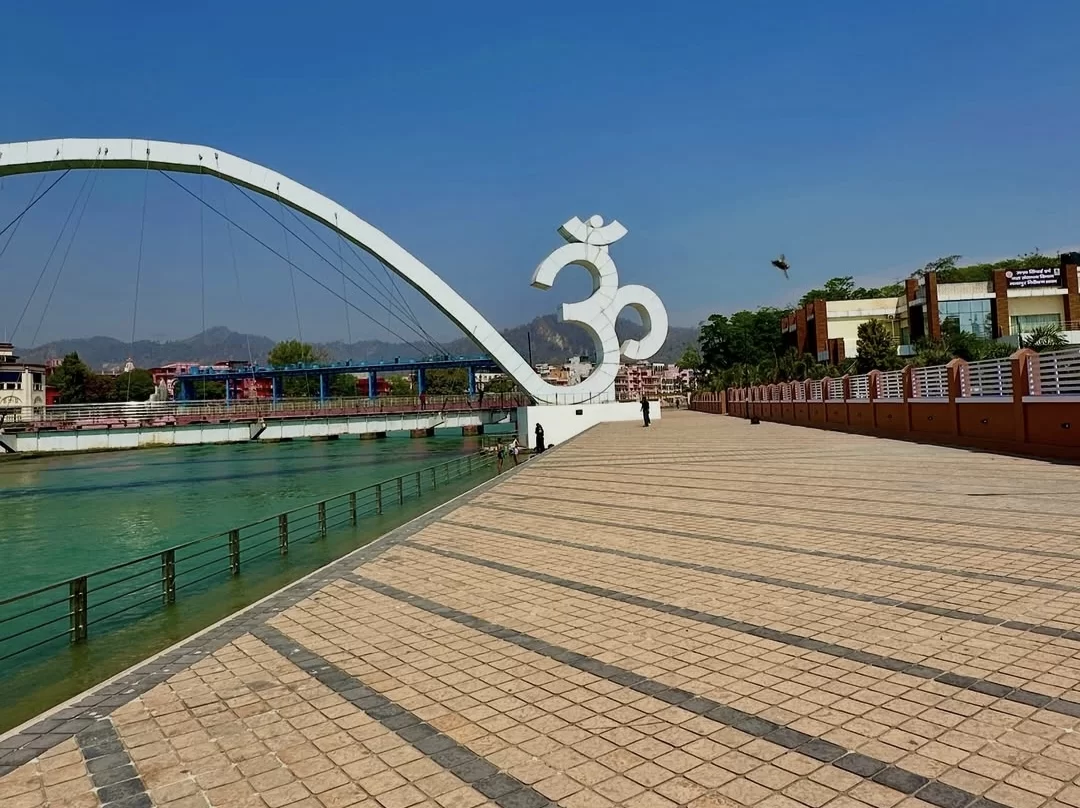 Har Ki Pauri Haridwar iconic white Om symbol on modern curved pedestrian bridge over Ganga river mountains blue sky green trees, perfect spiritual riverside walk, Uttarakhand tour packages.