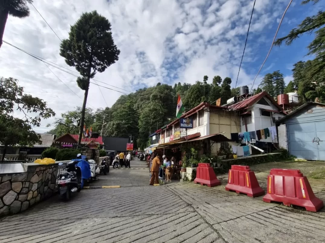 Landour Bazaar street during sunny day, featuring shops, scooters, Indian flag and deodar backdrop, perfect adventure Uttarakhand tour package.