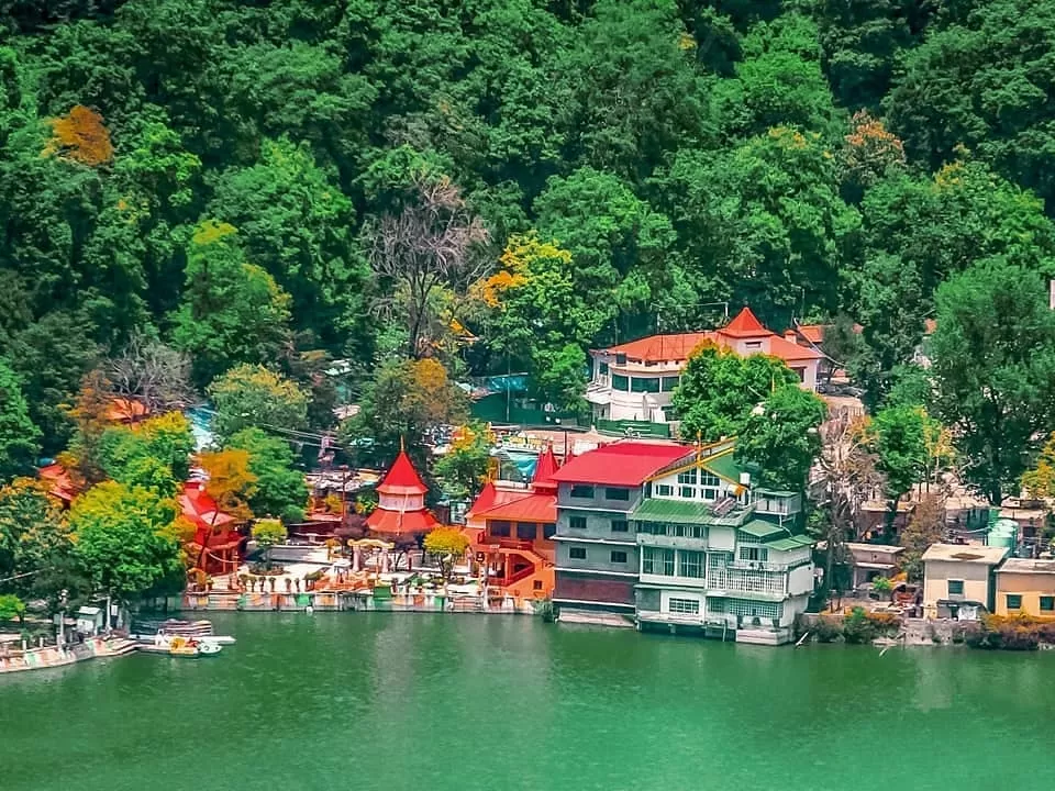 Naina Devi Temple panoramic aerial view overlooking Naini Lake Nainital Uttarakhand, showcasing cluster of red conical roof temples resorts amid lush green autumn foliage hills reflected in emerald waters with boats, perfect Uttarakhand tour packages.