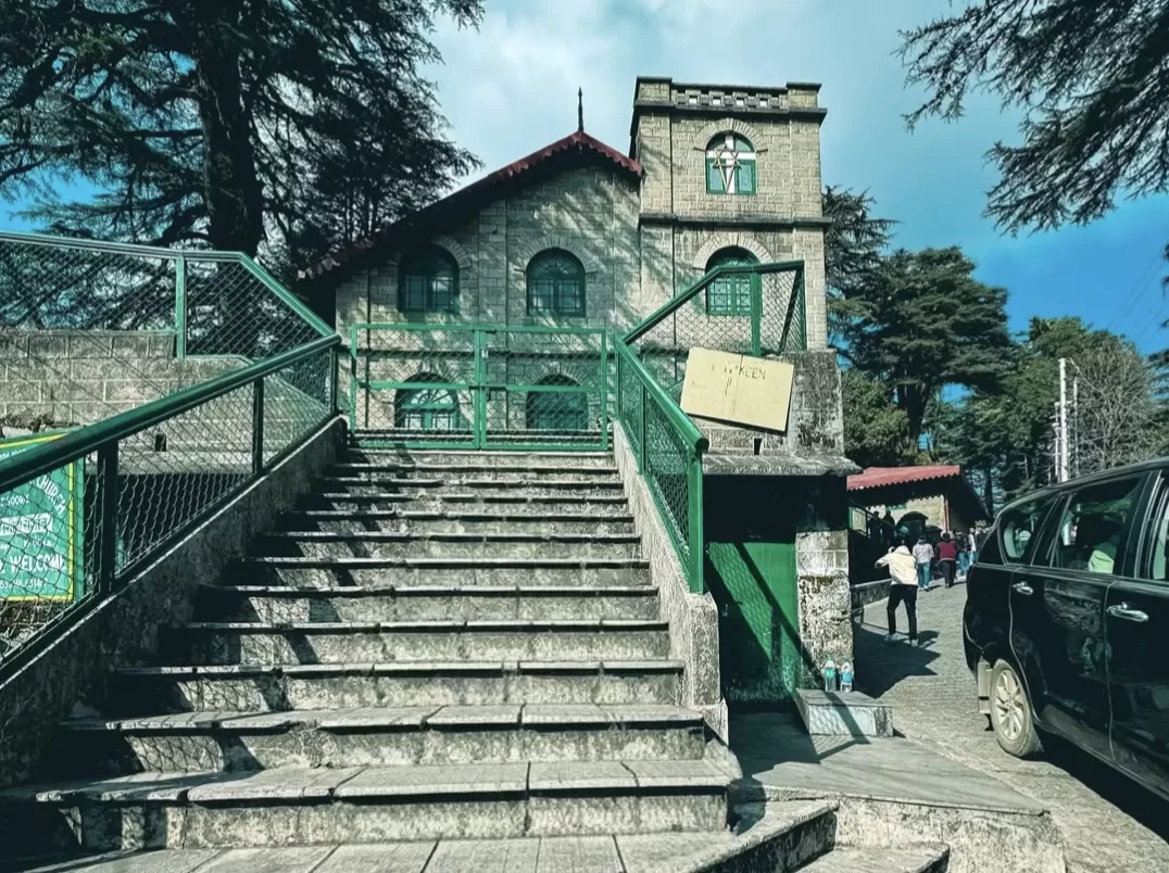 St Paul's Church entrance at Landour during partly cloudy day, featuring stone tower, green railings, stairs and deodar trees, perfect romantic Mussoorie tour package.