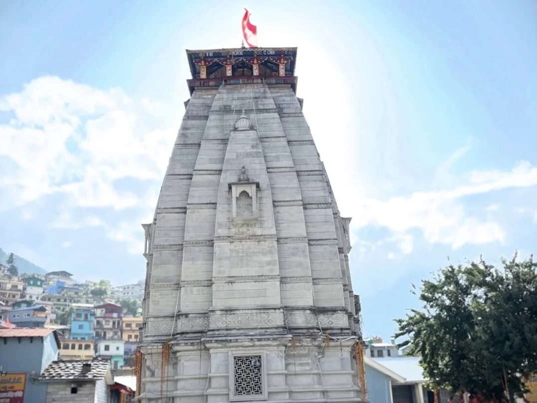 Narsingh Temple Joshimath soaring grey stone shikhara red flag golden roof accents amidst colorful hillside village homes green trees distant mountains blue sunny skies scattered clouds, perfect Uttarakhand tour packages.
