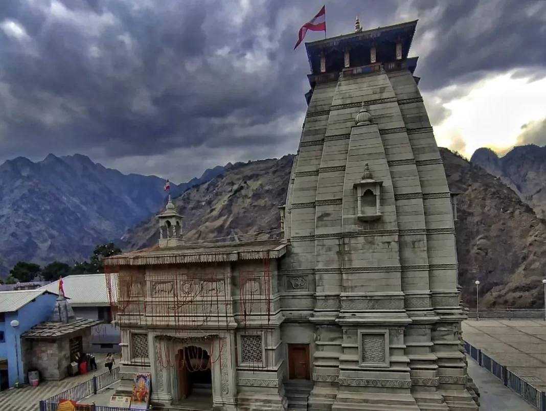 Narsingh Temple Joshimath dramatic grey stone pagoda tower red flags under brooding dark clouds nestled against rugged brown Himalayan mountains colorful village buildings foreground, perfect Uttarakhand tour packages.