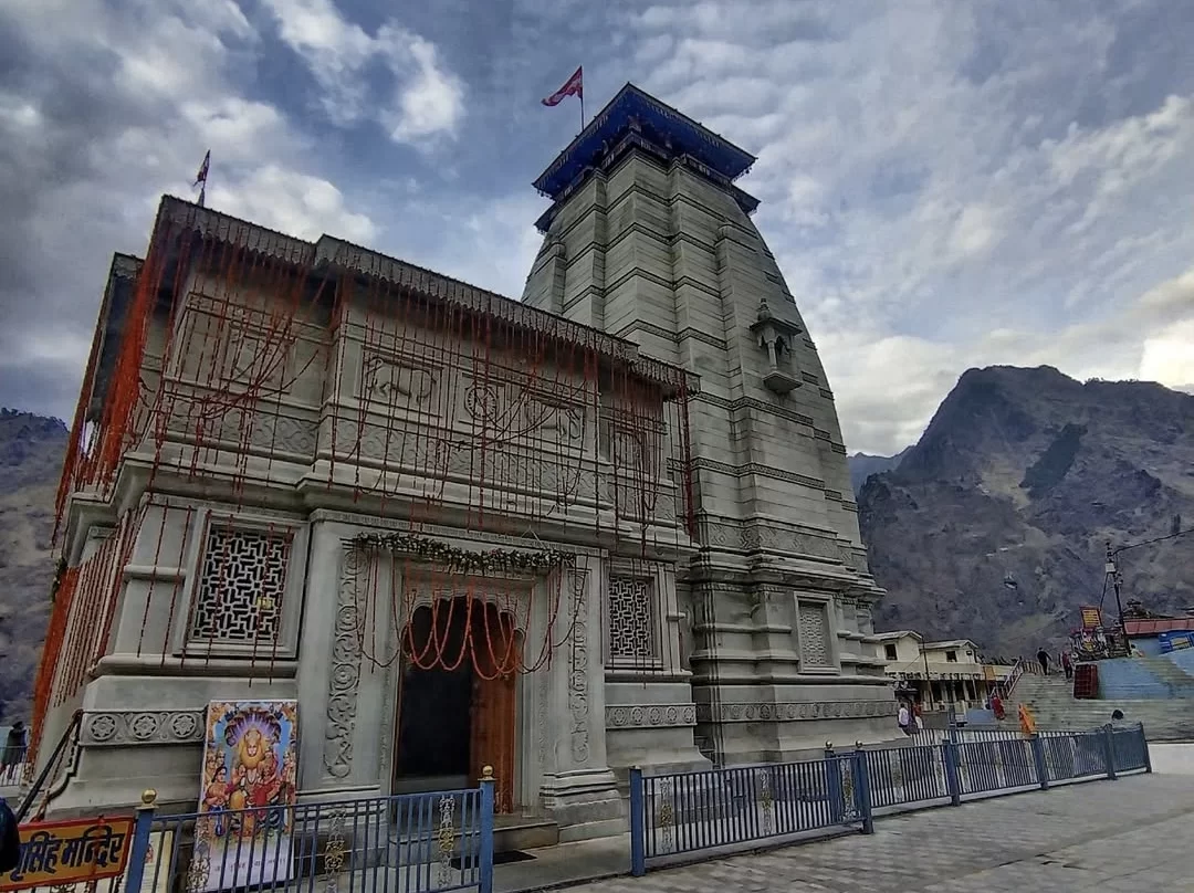 Narsingh Temple Joshimath elegant grey stone temple tiered tower red flag orange garlands lattice screens ornate entrance Lakshmi poster blue railing paved courtyard dramatic cloudy skies mountain backdrop, perfect Uttarakhand tour packages.