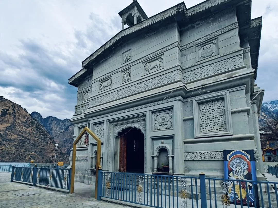 Narsingh Temple Joshimath grand grey stone facade elaborate lattice carvings lion motifs arched golden torana entrance Vishnu poster blue fence paved courtyard rugged mountain backdrop cloudy skies, perfect Uttarakhand tour packages.