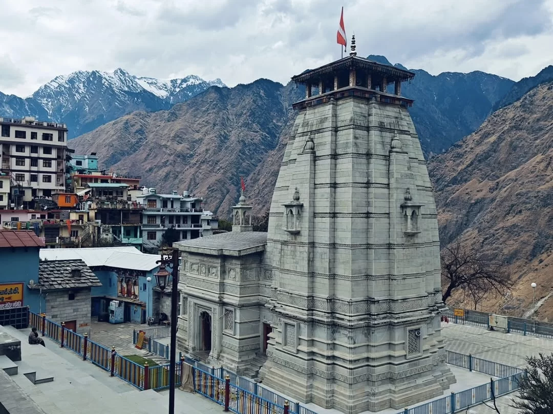 Narsingh Temple Joshimath panoramic view grey stone pagoda tower red flags amidst colorful clustered hillside buildings against rugged snow-capped Himalayan peaks cloudy skies, perfect Uttarakhand tour packages.