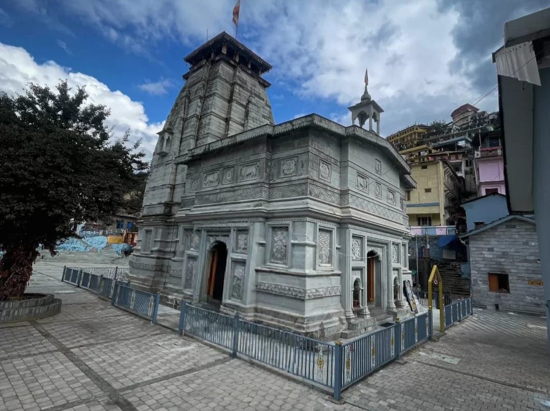 Narsingh Temple Joshimath ornate grey stone temple multi-tiered shikhara red flags surrounded by colorful hillside buildings trees blue metal fence paved courtyard cloudy skies, perfect Uttarakhand tour packages.
