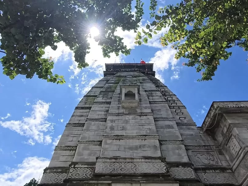 Narsingh Temple Joshimath majestic stone shikhara towering upward intricate carvings red flag sun rays filtering through lush green leaves blue skies scattered clouds, perfect Uttarakhand tour packages.