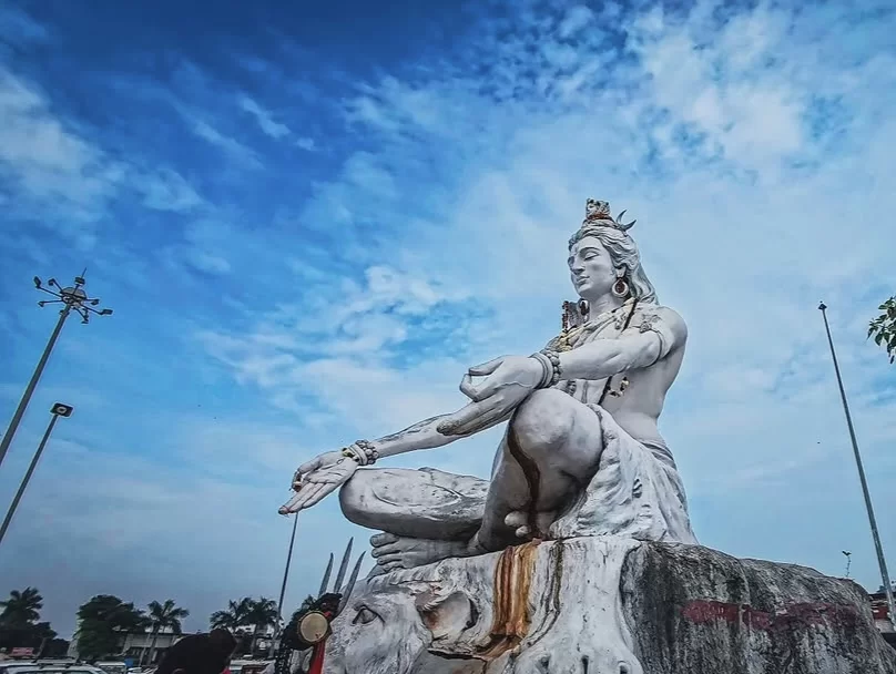 Majestic white marble Lord Shiva statue seated in meditative pose at Narsingh Temple Joshimath, blue skies with clouds, palm trees, street lamps, people nearby, perfect Uttarakhand spiritual tour package.