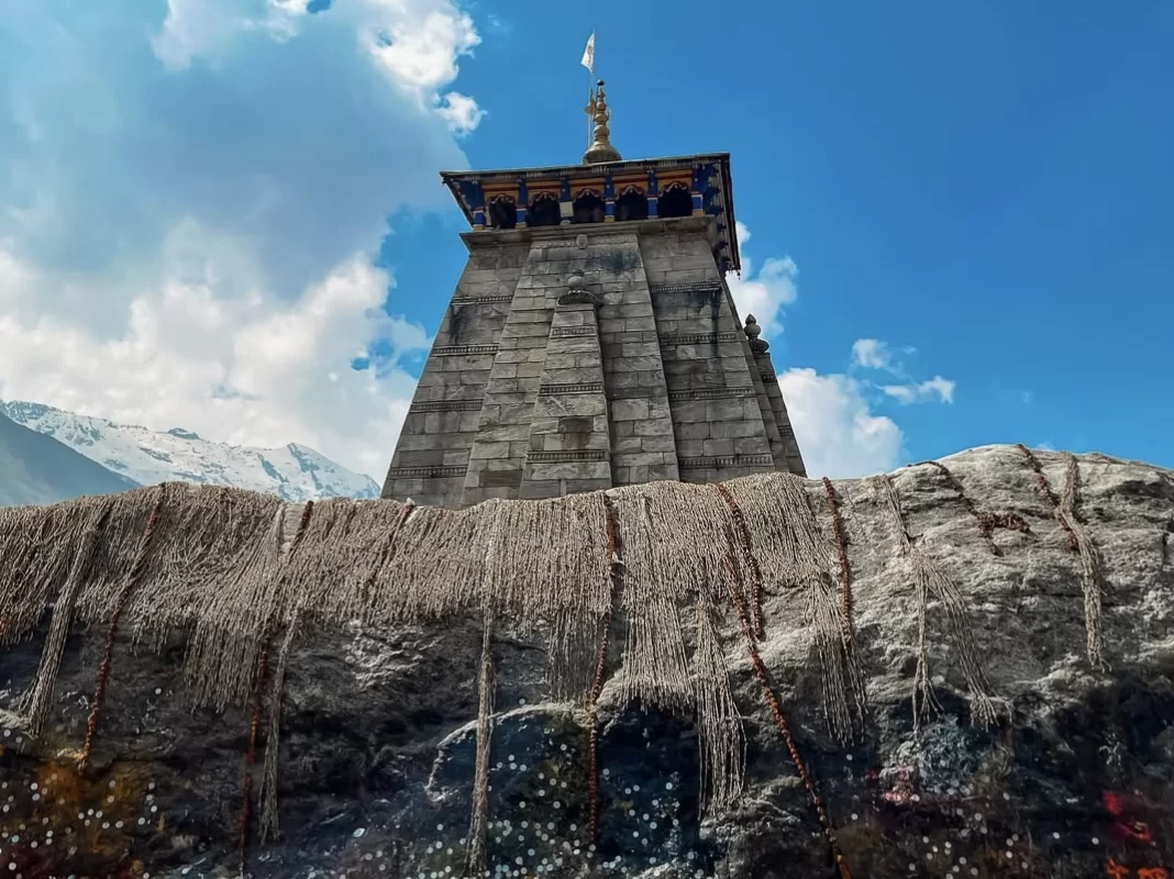 Iconic grey shikharas of Kedarnath Temple at Kedarnath during sunny day, featuring snowy peaks orange flags blue skies rocky foreground, perfect spiritual Uttarakhand tour packages.