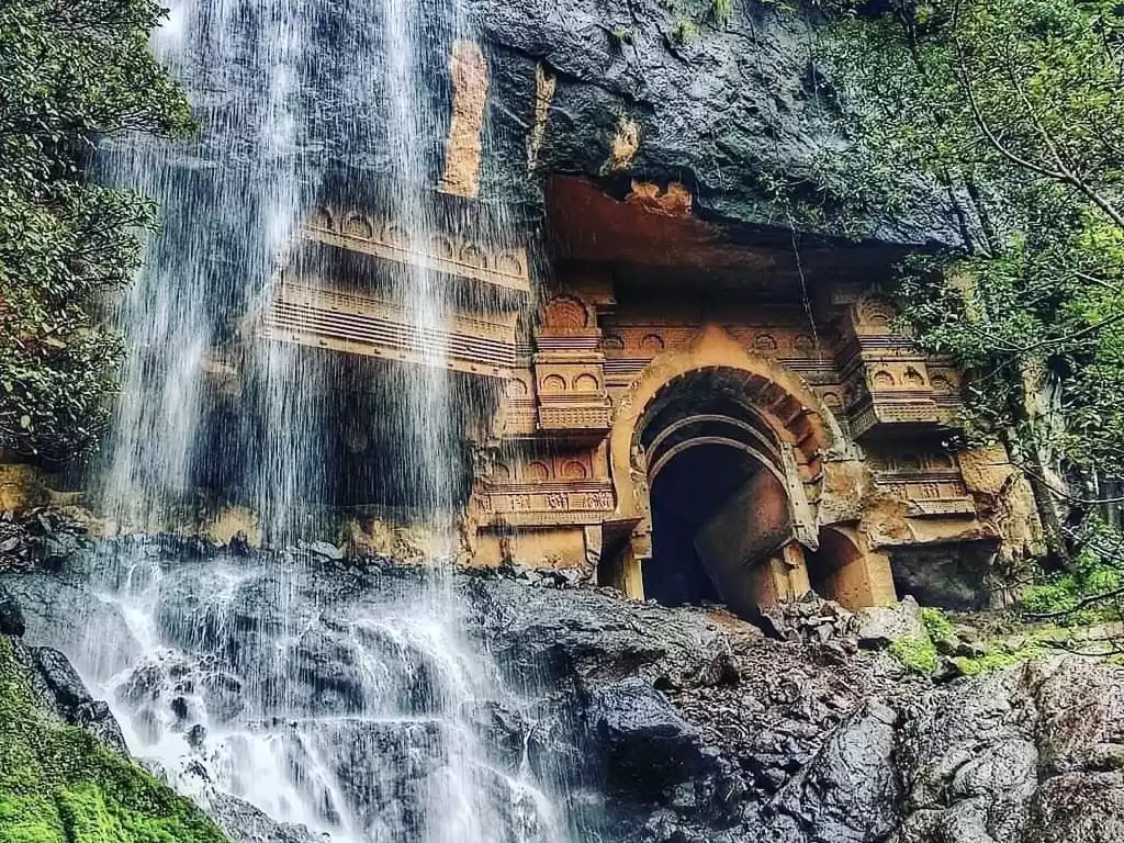 Ancient rock cut chaitya facade with cascading waterfall at Karla Caves Maharashtra during monsoon, featuring intricate carvings greenery cliff, perfect heritage experience Maharashtra cave tour package.
