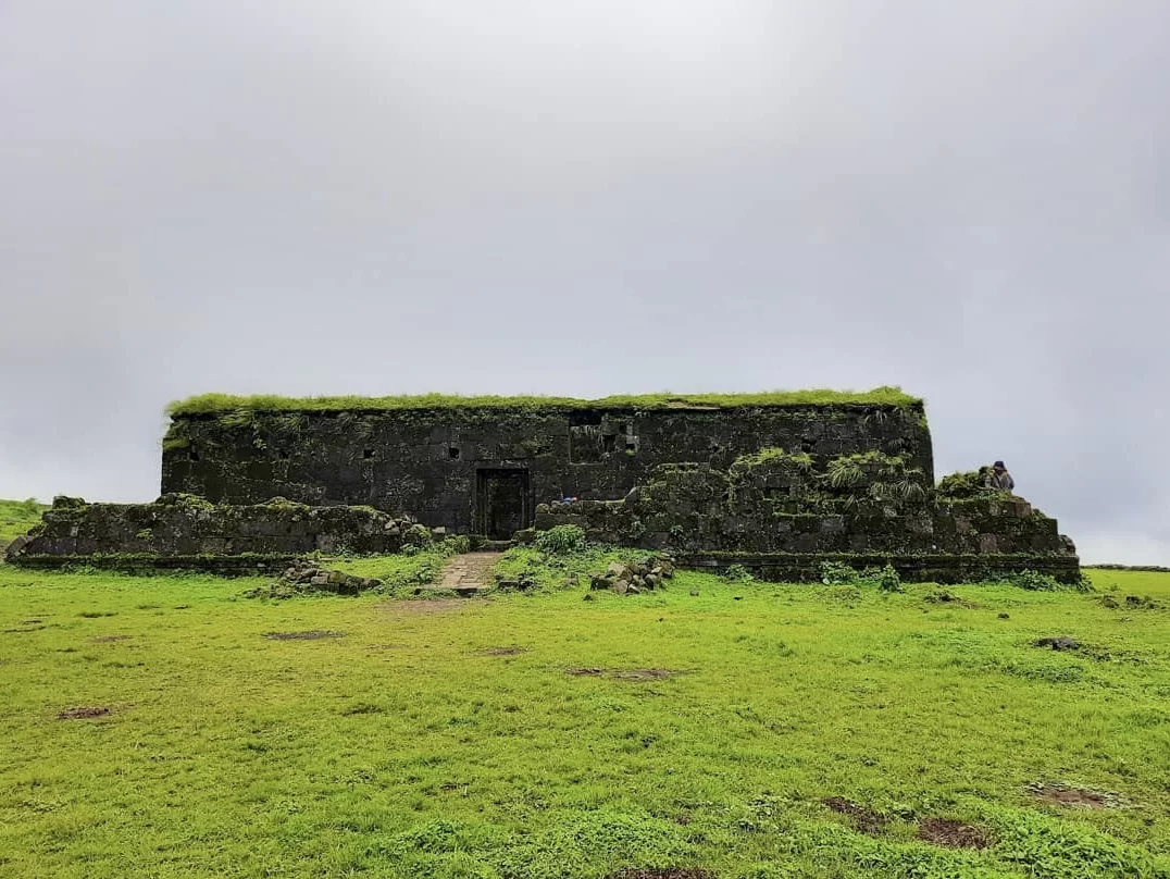Ancient ruins at Visapur Fort during overcast monsoon, featuring moss-covered walls & green grasslands, perfect adventure experience Maharashtra tour packages.