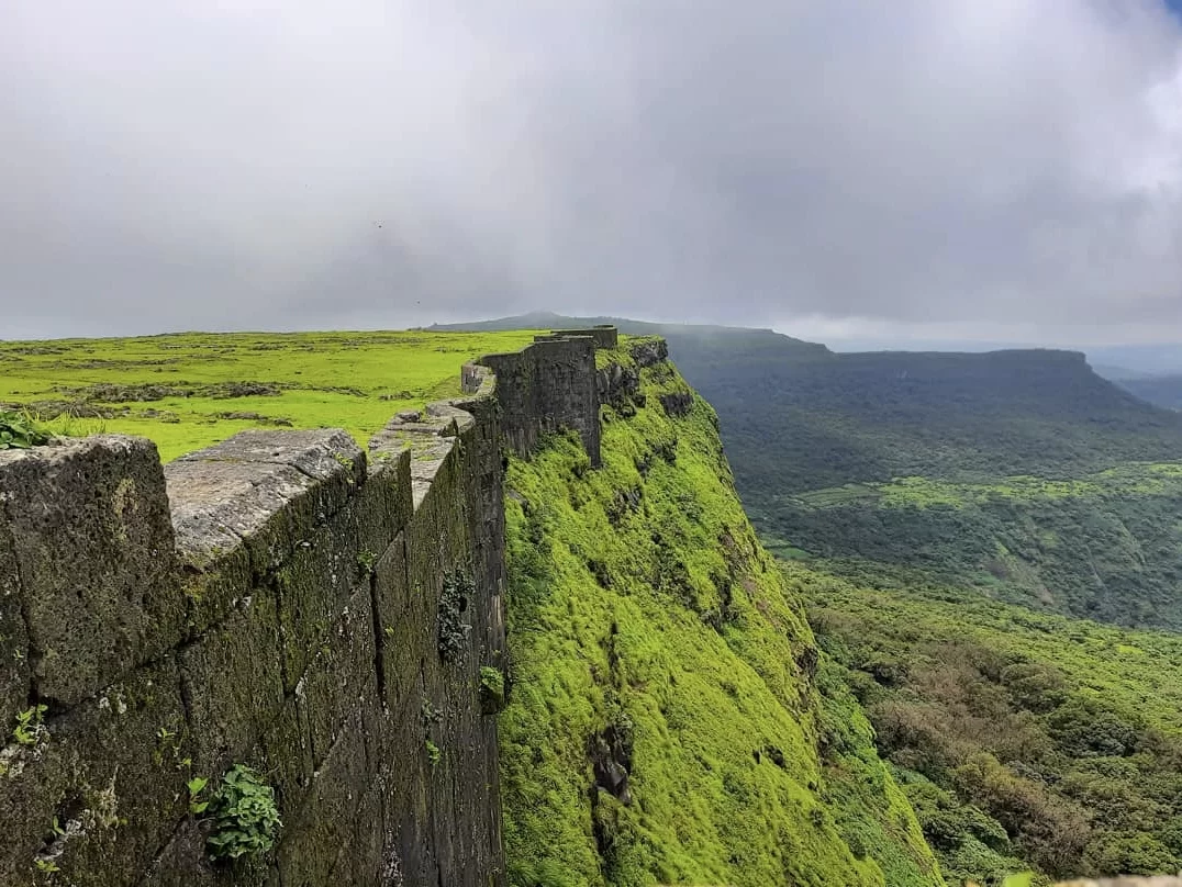 Cliff edge at Visapur Fort during misty monsoon, featuring green plateau & valley views, perfect adventure experience Maharashtra tour packages.