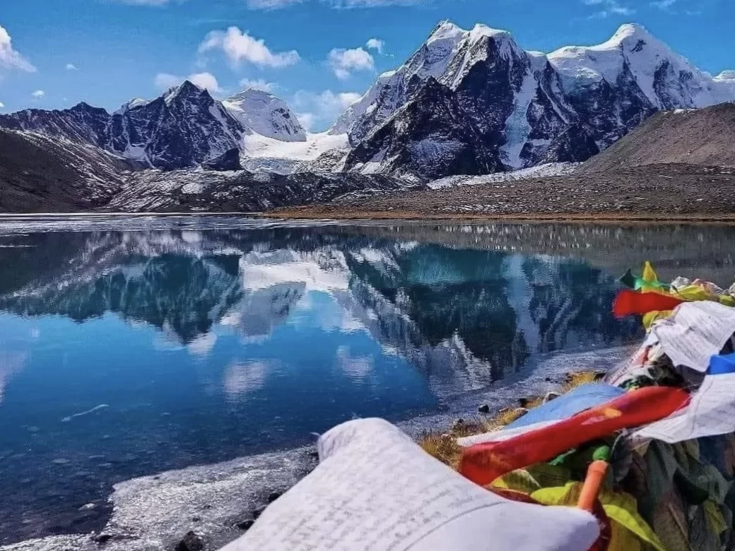 Gurudongmar Lake panoramic view at North Sikkim during sunny day, featuring turquoise waters snow peaks rocky banks clouds, perfect high-altitude adventure Sikkim tour package.