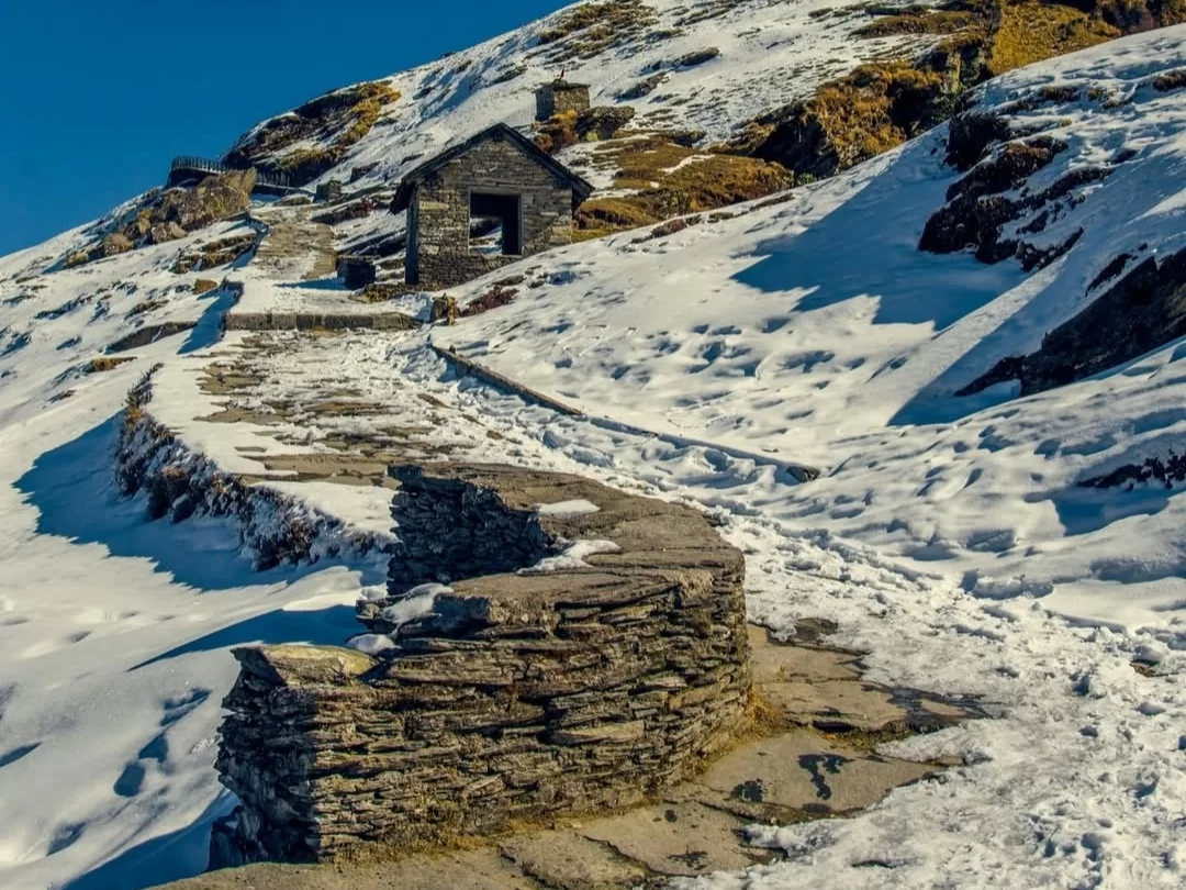 Chandrashila Peak winding snowy stone pathway curving uphill small stone hut pine trees snowy rocky Himalayan slopes blue sky backdrop, perfect Tungnath summit trek panoramic views Chopta Uttarakhand Tour Packages.