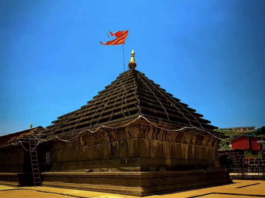 Mahabaleshwar Temple shikhara during sunny day Maharashtra, featuring tiered black stone spire saffron flag gold kalash clear blue sky hill backdrop, perfect hill station tour packages.