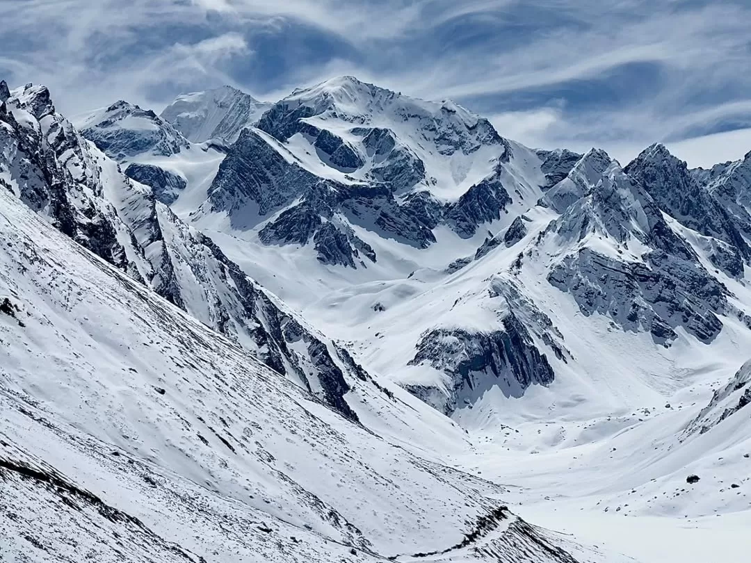 Om Parvat soaring snow summit amid swirling clouds and vivid blue skies, highlighting bold black rock spines and sweeping glacial fields encircled by towering Himalayan neighbors, perfect Uttarakhand tour package.