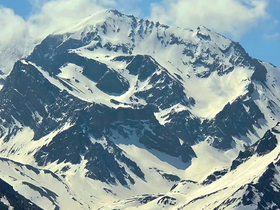 Om Parvat majestic snow summit piercing parted clouds under brilliant blue sky, revealing sheer dark rock faces and vast glacial aprons flanked by rugged Himalayan ridges, perfect Uttarakhand tour package.