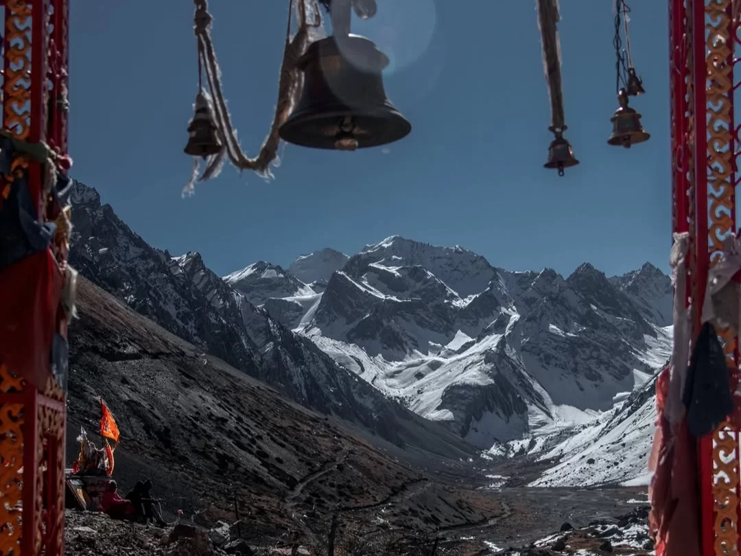 Om Parvat framed through ornate temple toran with dangling brass bells, prayer scarves and lit diya, showcasing snow-clad peaks in valley under clear blue sky, perfect Uttarakhand tour package.