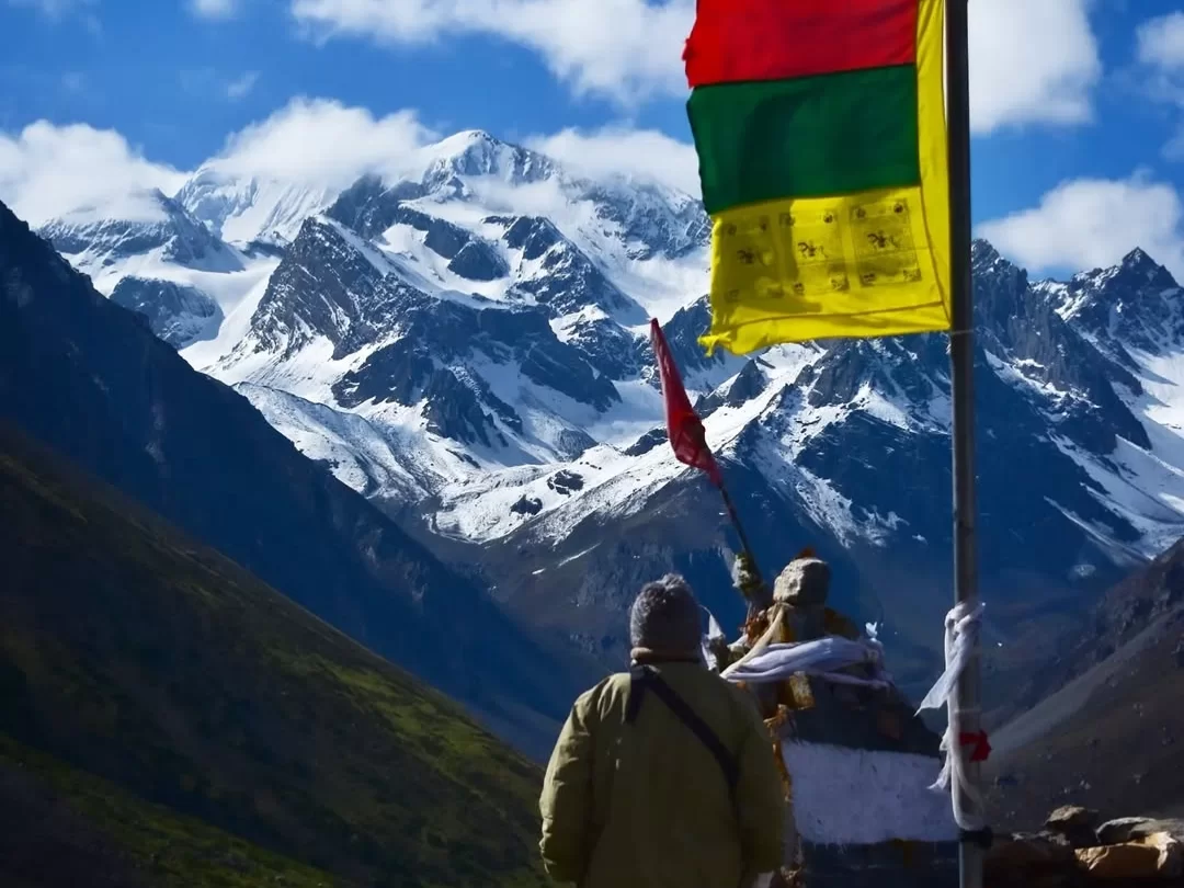 Pilgrims offering prayers with prayer flags fluttering in front of Om Parvat under partly cloudy blue skies, featuring snow-covered Himalayan peaks, green valley backdrop and traditional Tibetan scarves, perfect Uttarakhand tour package.