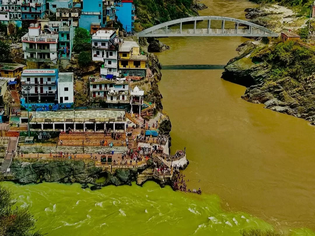 Devprayag Uttarakhand riverside colorful blue buildings crowded ghats suspension bridge brown Alaknanda River rocky banks green hills, perfect Uttarakhand tour packages.