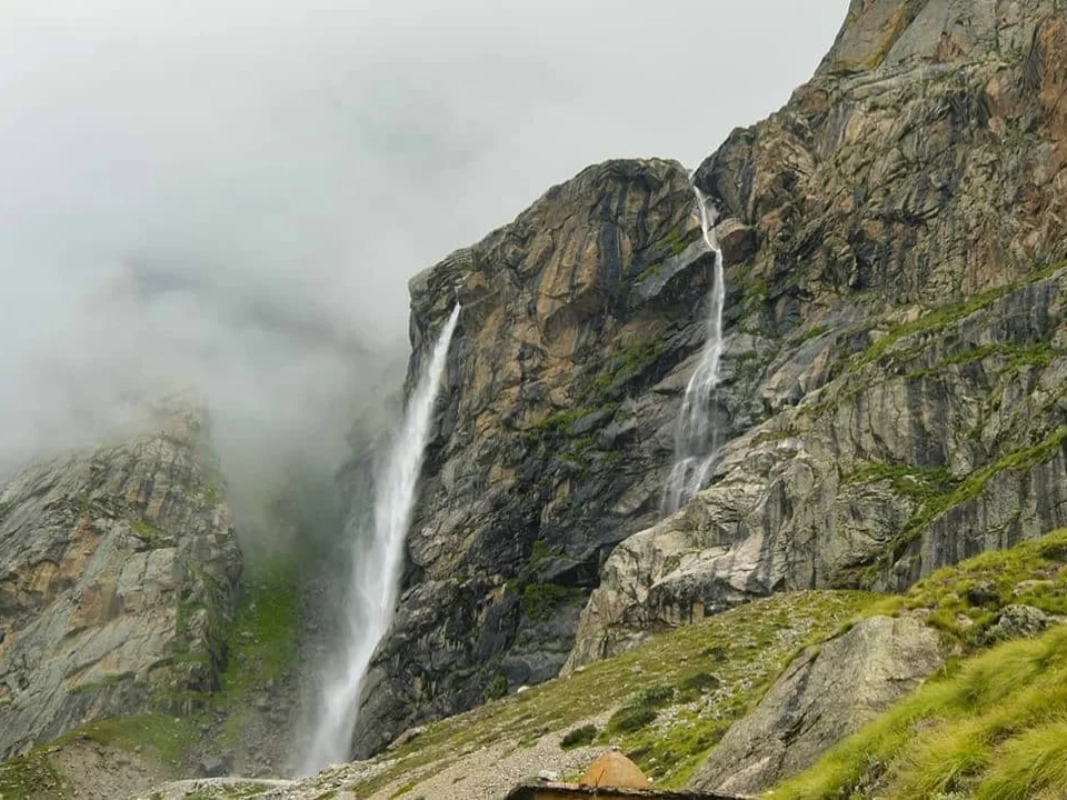 Vasudhara Falls Badrinath multiple misty waterfalls steep rocky cliffs green meadows cloudy sky Uttarakhand Char Dham trekking tour packages. 