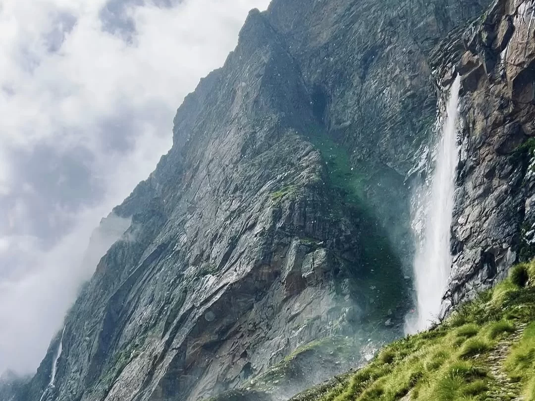 Vasudhara Falls Badrinath cloudy sky tall waterfall steep rocky cliffs green grassy slopes trekking path Uttarakhand Char Dham tour packages.