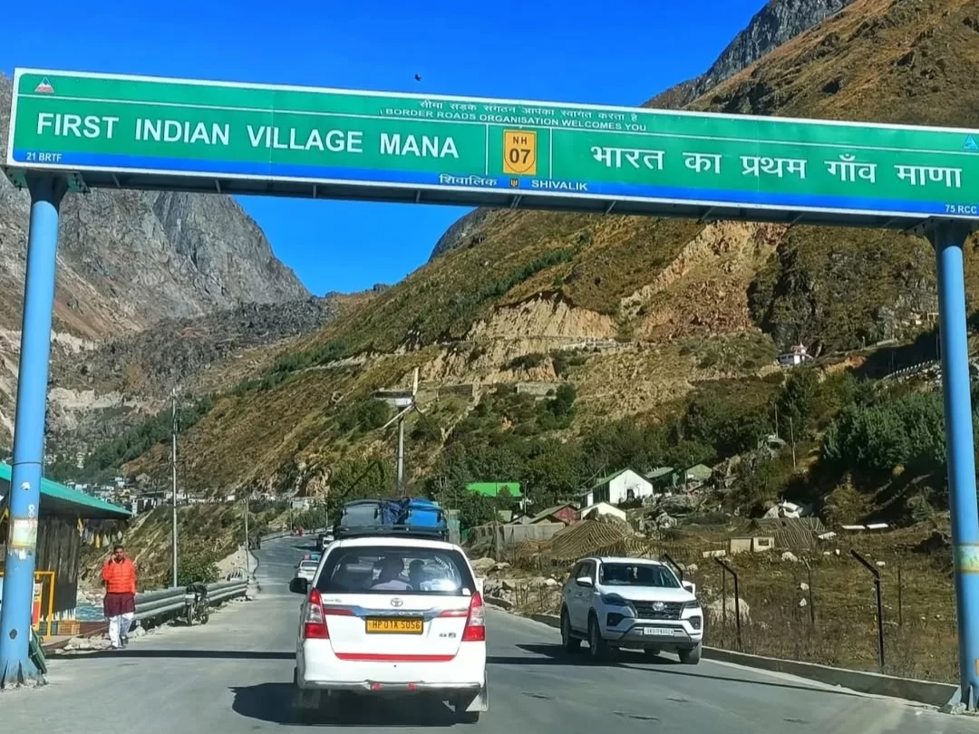 Mana Village First Indian Village signboard blue arch Hindi English Badrinath road snowy mountains vehicles tourists Uttarakhand tour packages. 