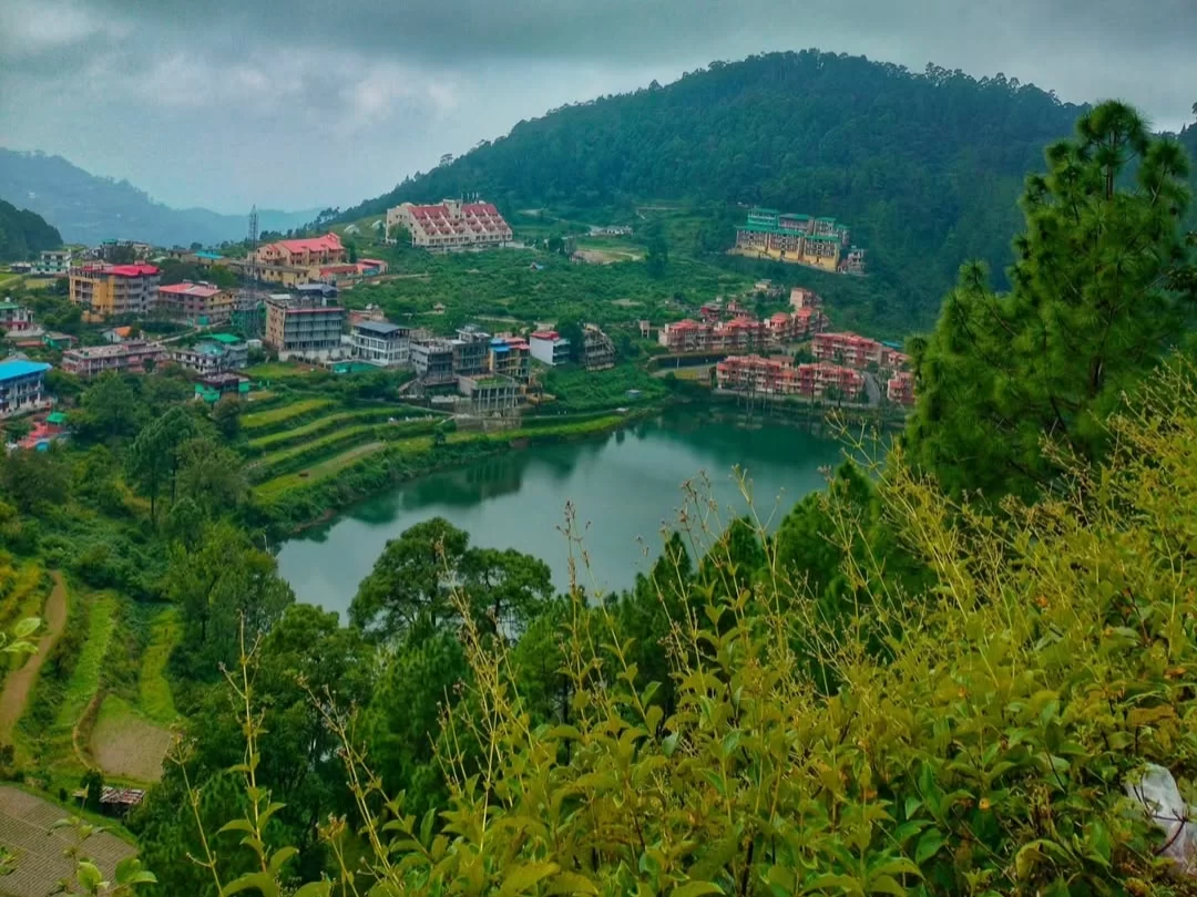 Khurpa Tal Nainital scenic lake view during cloudy day, featuring colorful buildings terraced fields surrounding green hills pine trees, perfect nature romantic experience Uttarakhand tour packages.