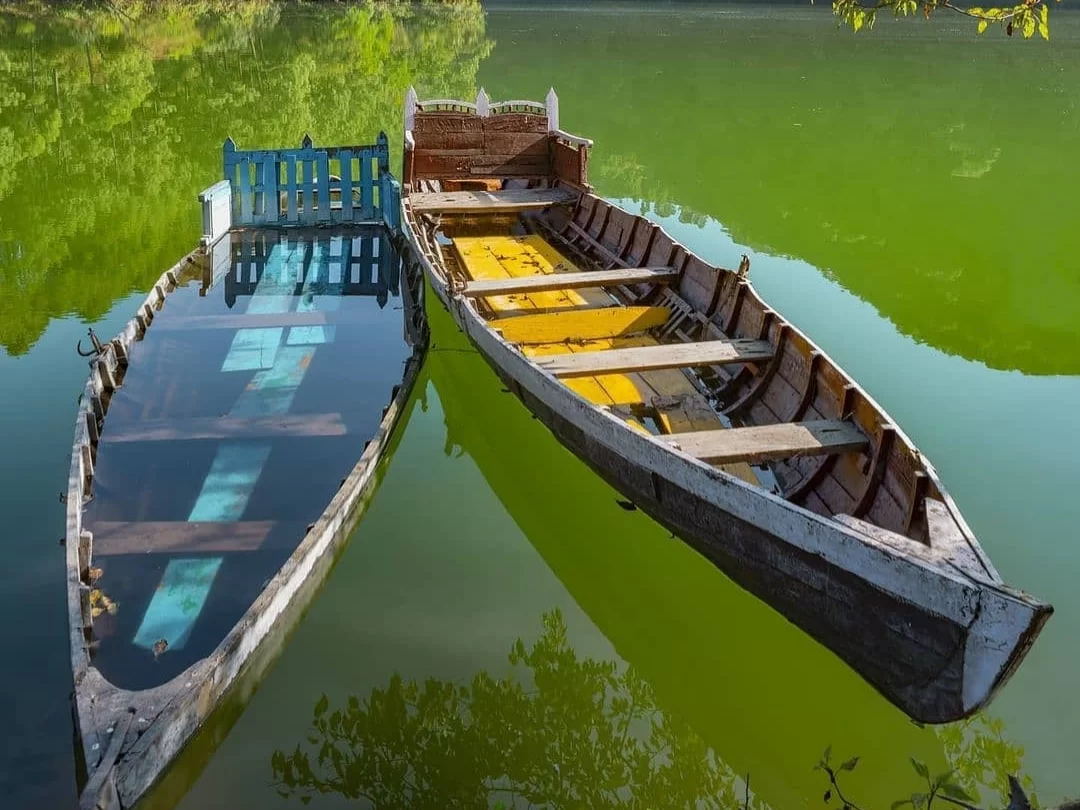 Sattal Lake Nainital traditional wooden rowboat yellow stripes blue railing dock reflected emerald green calm waters lush green foliage backdrop, perfect Uttarakhand tour packages.