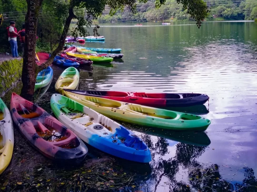 Sattal Lake Nainital colorful kayaks red orange green blue lined shore lush green trees person standing serene reflective lake water distant boat Uttarakhand tour packages.