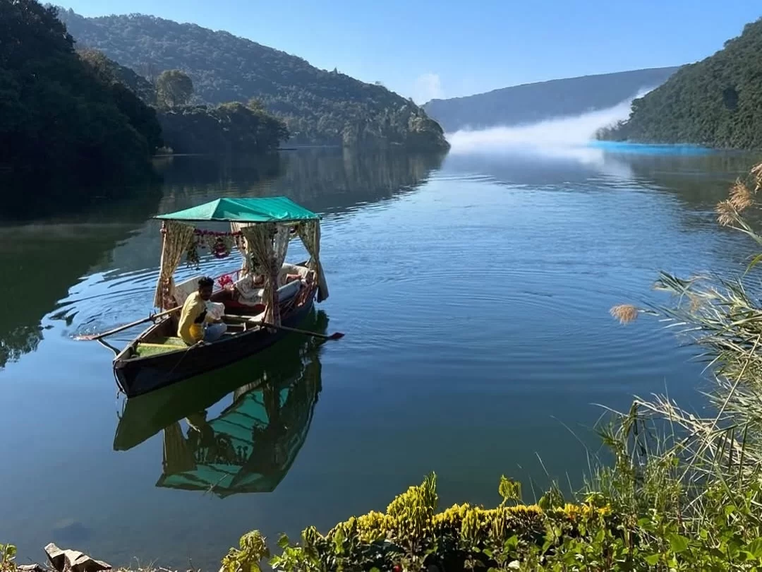 Shikara boat ride on Naukuchiatal Lake Nainital during misty sunny morning, featuring green canopy, passengers and forested mountains, perfect romantic experience Uttarakhand tour package.