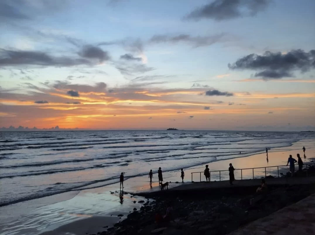 Silhouettes on Varsoli Beach in Alibaug at sunset, featuring orange sky, waves and pier with people, perfect Alibaug tour package.