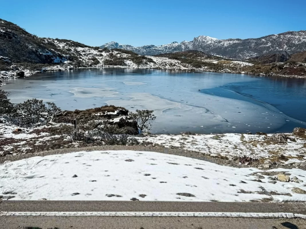 Bum La Pass Tawang Arunachal Pradesh winter high altitude frozen lake snowy rugged mountains blue water ice edges bushes road blue sky clouds, perfect Arunachal tour packages.