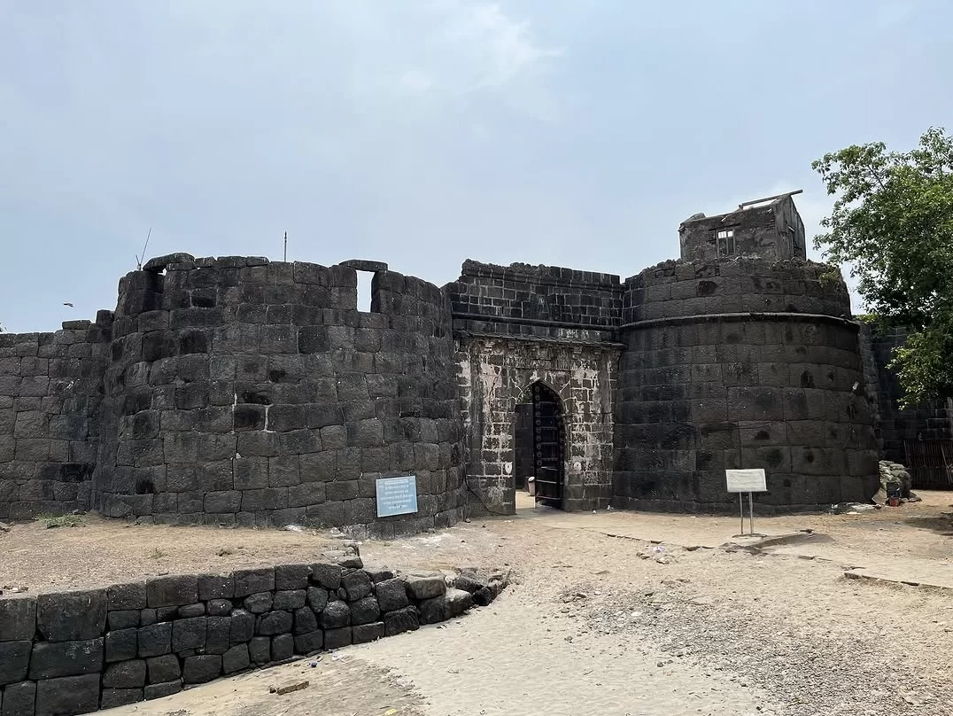 Ground-level view of Kolaba Fort’s massive black stone walls and arched entrance gate, with rounded bastions, weathered masonry, and open sandy foreground under a clear sky.