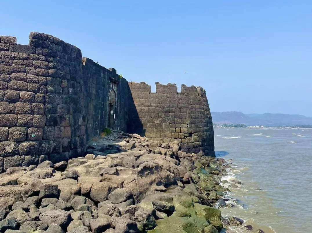 Seaside stone ramparts of Kolaba Fort rising directly from the rocky shoreline, with waves lapping at the base of the weathered black basalt walls and distant hills visible across the water.
