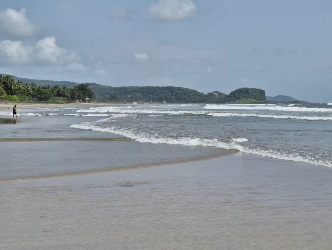 Solitary figure strolling Kashid Beach during partly cloudy daytime, featuring wide grey sands, rolling waves, lush green hills backdrop, perfect serene beach walk experience with Konkan coast tour package