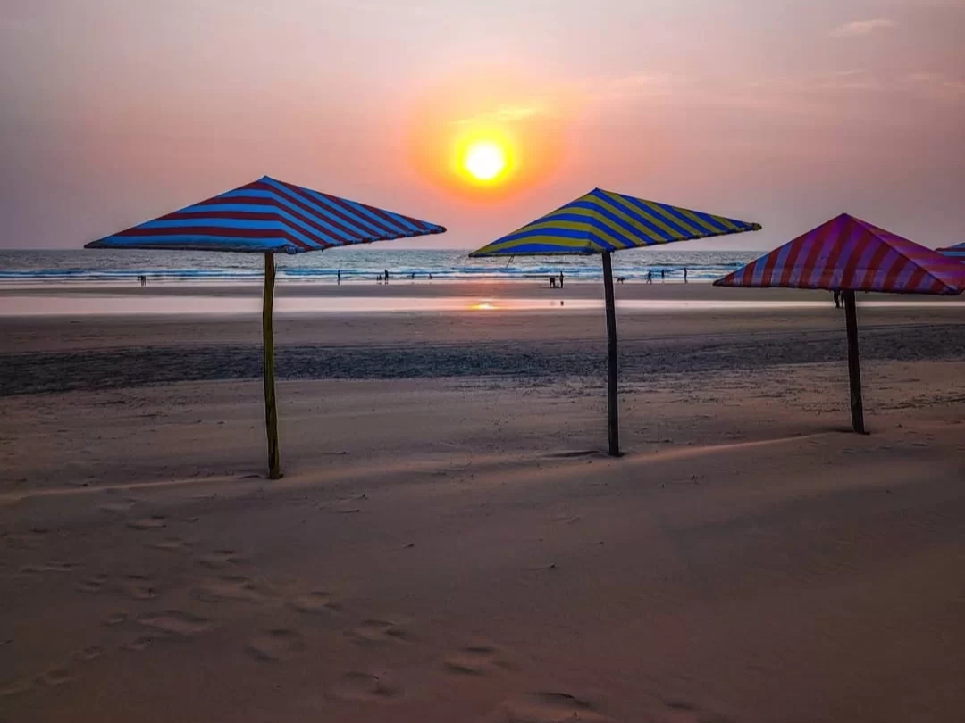 Golden hour silhouette of colorful striped beach umbrellas on sandy shore Mandrem Beach North Goa with setting sun Arabian Sea waves distant people, perfect romantic sunset experience with Mandrem Beach Goa tour package.