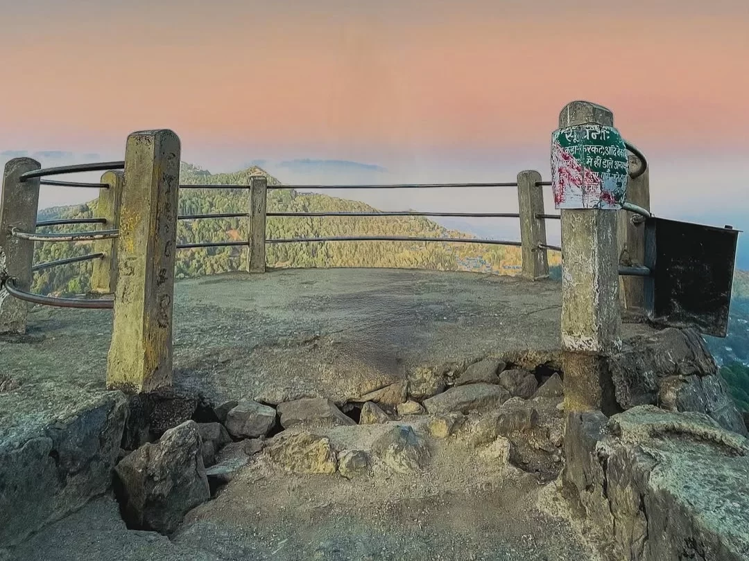Tiffin Top Nainital weathered signpost Tiffin Top green red notices on rusty railings rocky platform sunset pink orange skies pine hills terraced town backdrop, perfect Uttarakhand tour packages.