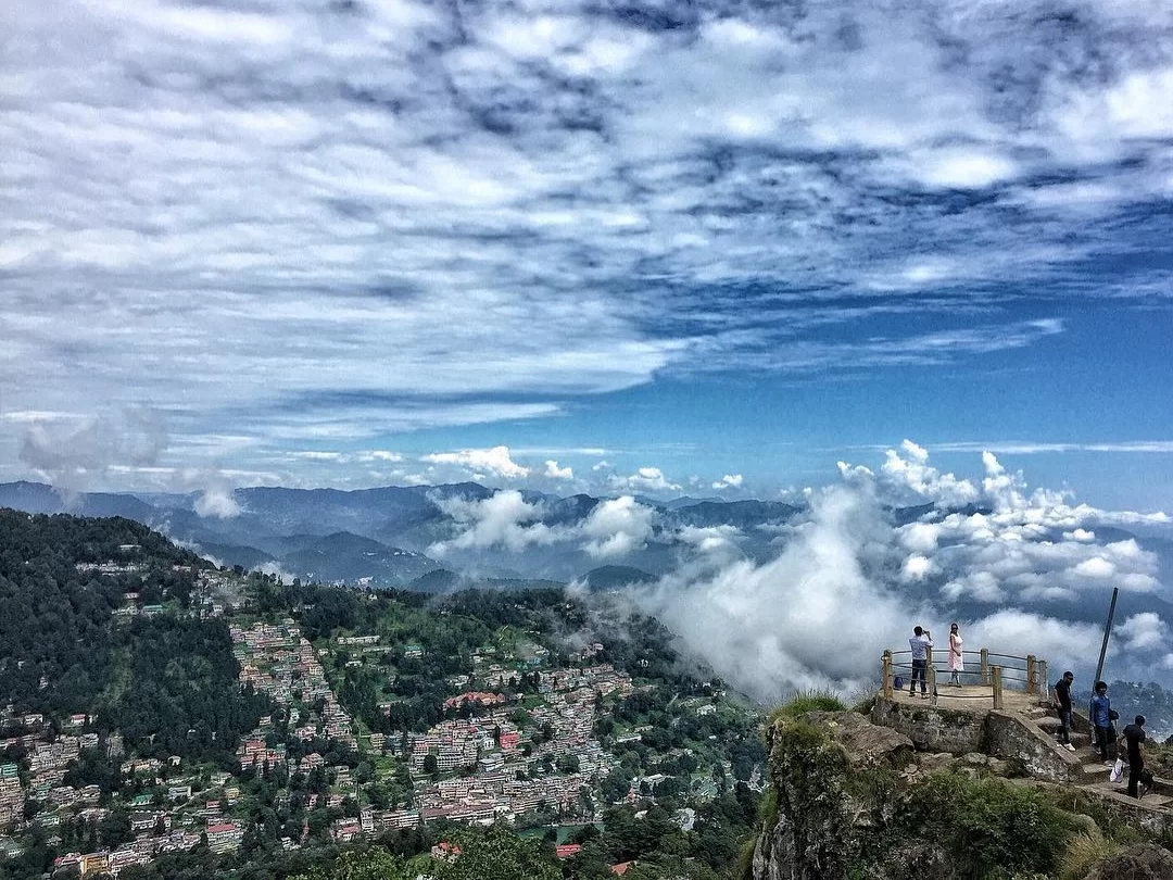 Tiffin Top Nainital elevated wooden viewpoint tourists gazing over mist-shrouded green pine hills terraced colorful town valley distant mountains dynamic blue skies scattered clouds, perfect Uttarakhand tour packages.
