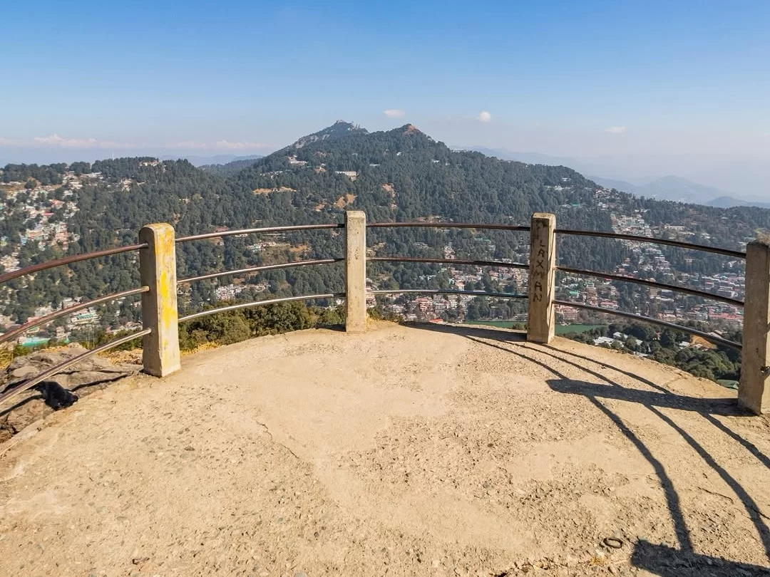 Tiffin Top Nainital elevated concrete viewpoint rusty yellow railings overlooking pine-clad hills terraced town distant snow peaks hazy blue skies clear weather, perfect Uttarakhand tour packages.