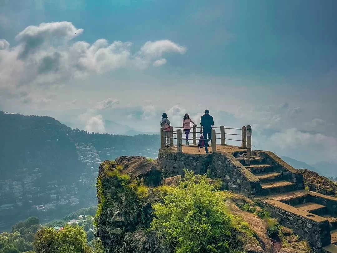 Tiffin Top Nainital cliffside stone viewing platform with tourists overlooking misty valley layered green hills terraced town blue skies fluffy clouds, perfect Uttarakhand tour packages.